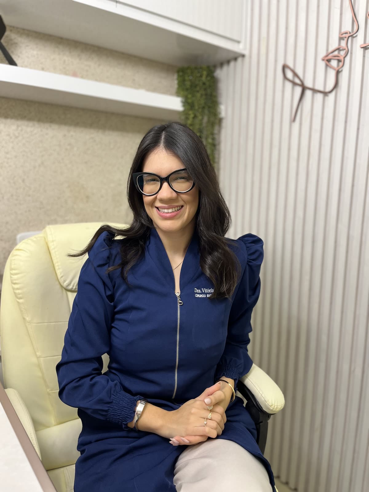 Dra. Vitória Machado sentada em seu consultório em Aracaju, sorrindo, com jaleco azul-escuro.